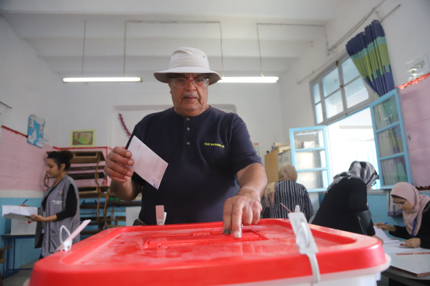 Ein Mann mit Hut stimmt in einer Wahllokal ab, vor einer roten Wahlurne stehend und ein Stück Papier in der Hand haltend, während andere an Tischen sitzen und schreiben, in der Nähe eines Fensters mit einem Vorhang.