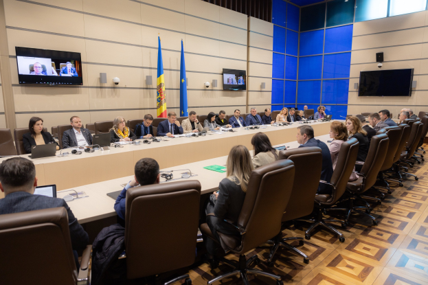 Große Gruppe von Menschen an einem langen Tisch mit Laptops, Mikrofonen, Papieren und anderen Gegenständen während einer Sitzung des Europäischen Rates für Menschenrechte, mit drei Flaggen, zwei Fernsehern und Deckenleuchten im Hintergrund.