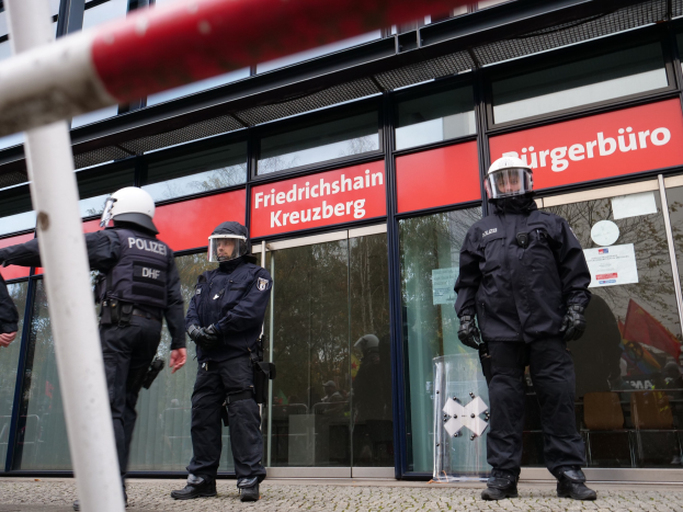 Polizisten in Uniform vor einem Glasgebäude mit aufgeklebten Papieren, neben einem Pfahl und Schildern mit der Aufschrift "Friedrichshain Kreuzberg."
