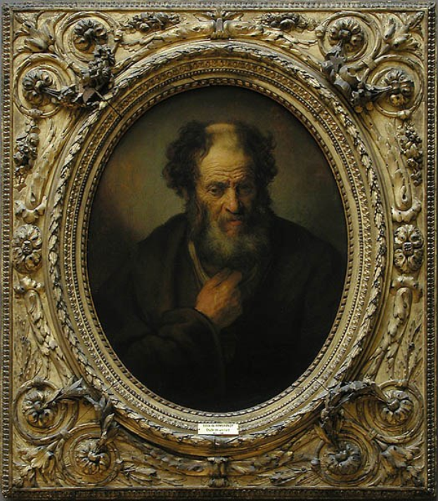 Portrait eines alten Mannes mit einem vollen, dunklen Bart und dunklem Haar, der eine weiße Robe trägt und mit gefalteten Händen in einem Raum mit weißen Wänden und einem Holzfußboden steht.