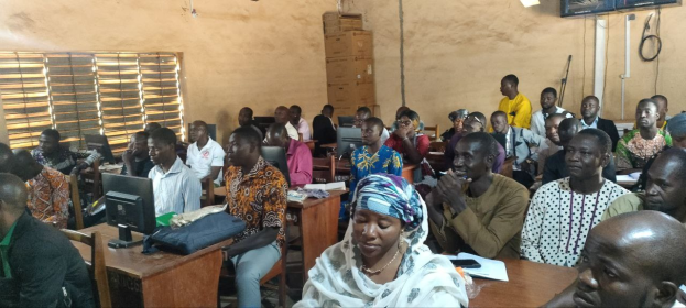 Eine Gruppe von Menschen sitzt an Tischen in einem Klassenzimmer mit Monitoren, Taschen, Büchern, Papieren und Kartons auf den Tischen, einem Fenster auf der linken Seite und einem Fernseher an der Wand im Hintergrund, an einer Schulung zum Thema Technologie teilnehmend.