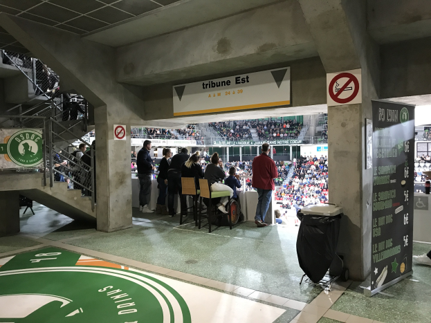 Eine Gruppe von Menschen steht auf einem Stadionfeld vor sitzenden Zuschauern, mit Schildern, die "Boston Celtics vs New York Jets" anzeigen, einem Müllcontainer, einer Treppe mit Geländern und Deckenleuchten.