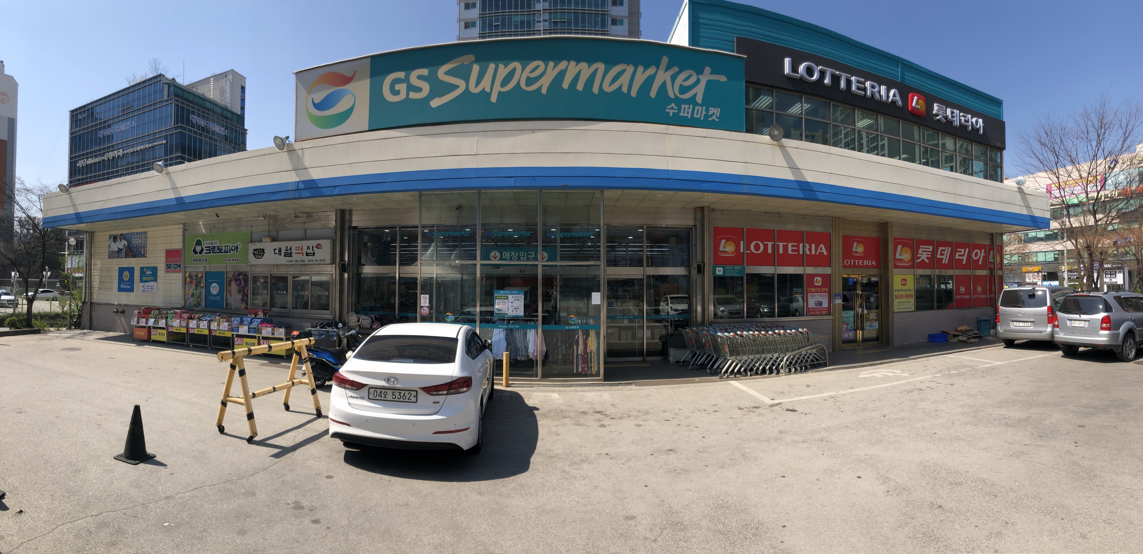 Ein Auto vor einem "GS Supermarkt"-Geschäft mit anderen Fahrzeugen, Verkehrskegeln und verstreuten Gegenständen auf der Straße, mit Gebäuden, Bäumen und einem klaren blauen Himmel im Hintergrund.