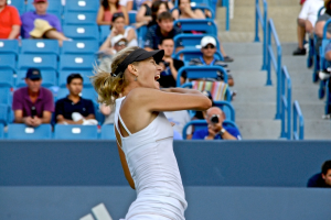 Eine Frau in weißem Oberteil und schwarzer Mütze steht vor einem blauen Stadionplatz mit Zuschauern.