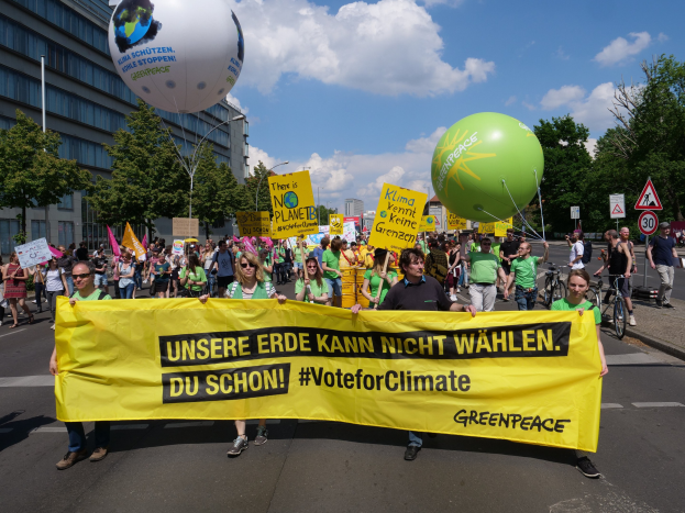Eine Gruppe von Menschen marschiert mit einem gelben "Wähle für das Klima"-Schild die Straße entlang, mit Ballons, Schildern, Fahrrädern, Bäumen, Laternenmasten und Gebäuden im Hintergrund unter einem klaren blauen Himmel.