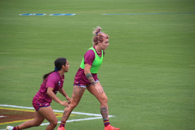 Zwei Frauen in Sportkleidung - eine in einem grünen T-Shirt und Shorts, die andere in einem pinken Kleid - spielen Fußball auf einem Feld und zeigen sichtbare Tattoos an Händen und Füßen.