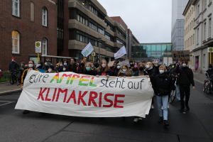 Eine Gruppe von Menschen marschiert mit einer "Klimakrise"-Plakette, Masken tragend, auf der Straße, mit Fahrrädern, Schildern und Gebäuden im Hintergrund unter einem klaren blauen Himmel.