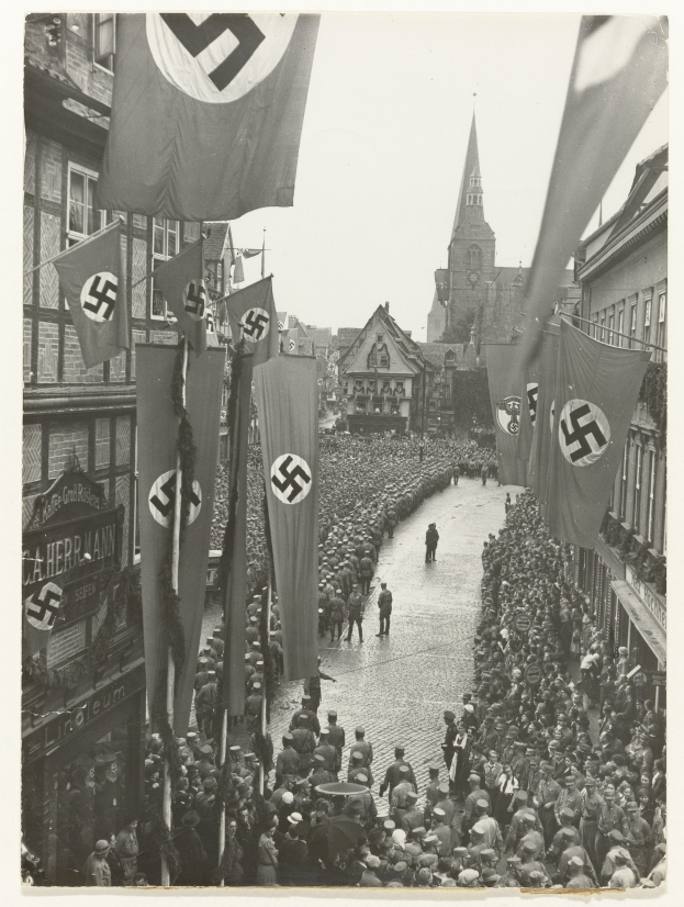 Schwarzes und wei├čes Foto einer gro├čen Menge, die mit Nazi-Flaggen die Stra├če entlangmarschiert, mit Geb├Ąuden und einem klaren Himmel im Hintergrund.