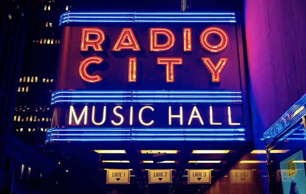 Radio City Music Hall in New York City, hell erleuchtet bei Nacht mit hohen Gebäuden in der Nähe, mit einer 'Radio City'-Leuchtschrift und zusätzlichen Schildern auf der rechten Seite.