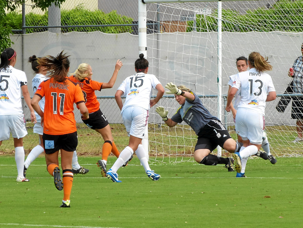 Eine Gruppe von Frauen spielt ein Spiel auf dem Boden mit einem Maschendrahtzaun und Bäumen im Hintergrund.