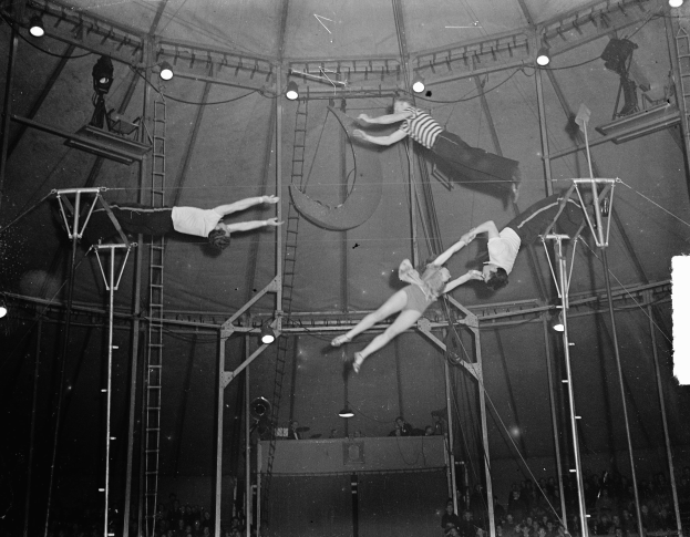 Ein Schwarz-Weiß-Foto einer Gruppe, die Artistik in einem Zirkuszelt performs, umgeben von Stangen, Leitern und Lichtern, mit einer Zuschauermenge darunter.