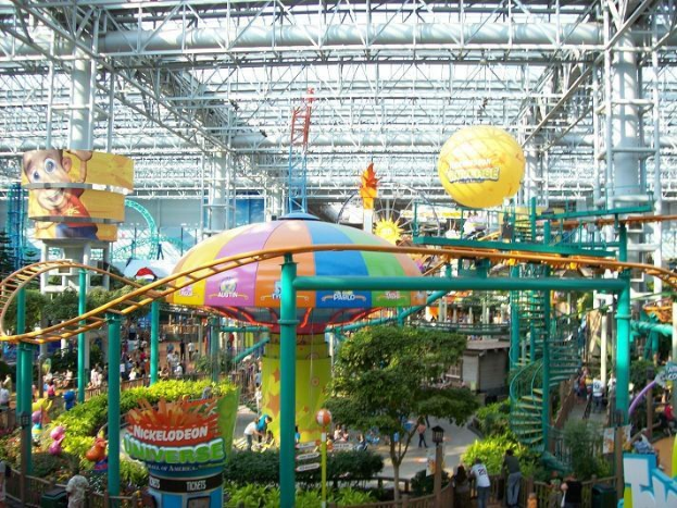 Großer Indoor-Wasserpark mit Achterbahn, umgeben von Menschen, Bäumen, Pflanzen und Deckenleuchten mit Stangen.