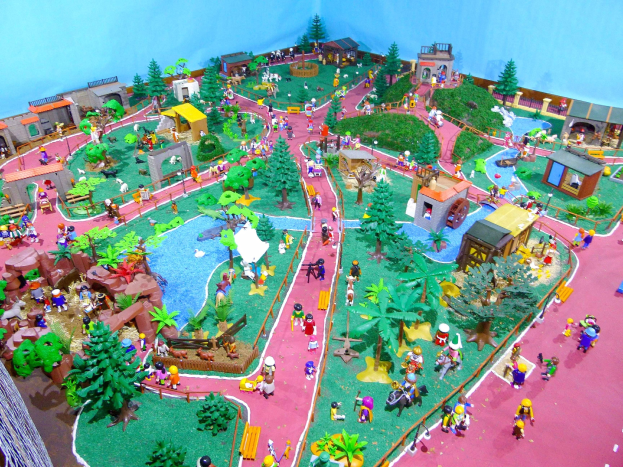 Zoo-Modell mit Miniaturfiguren, umgeben von Bäumen, Häusern, Pflanzen, Gras, einem Zaun und einer Hintergrundwand, mit Text unten.