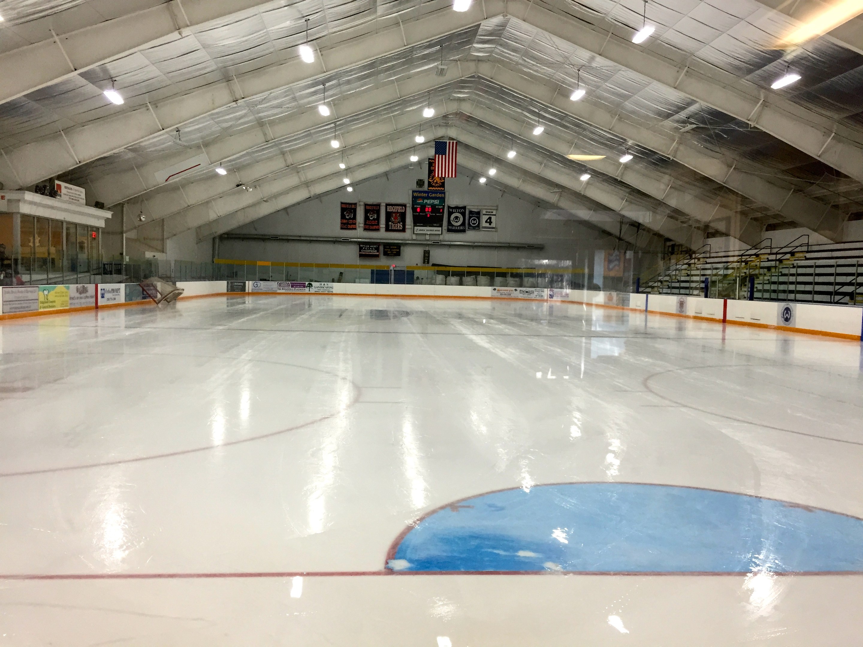 Großes Hallen-Eisstadion mit Deckenbeleuchtung, Werbetafeln, Treppen mit Geländern, einer Wand im Hintergrund und einigen Menschen, was darauf hinweist, dass es sich wahrscheinlich um eine Sportanlage handelt, die für Hockey genutzt wird.