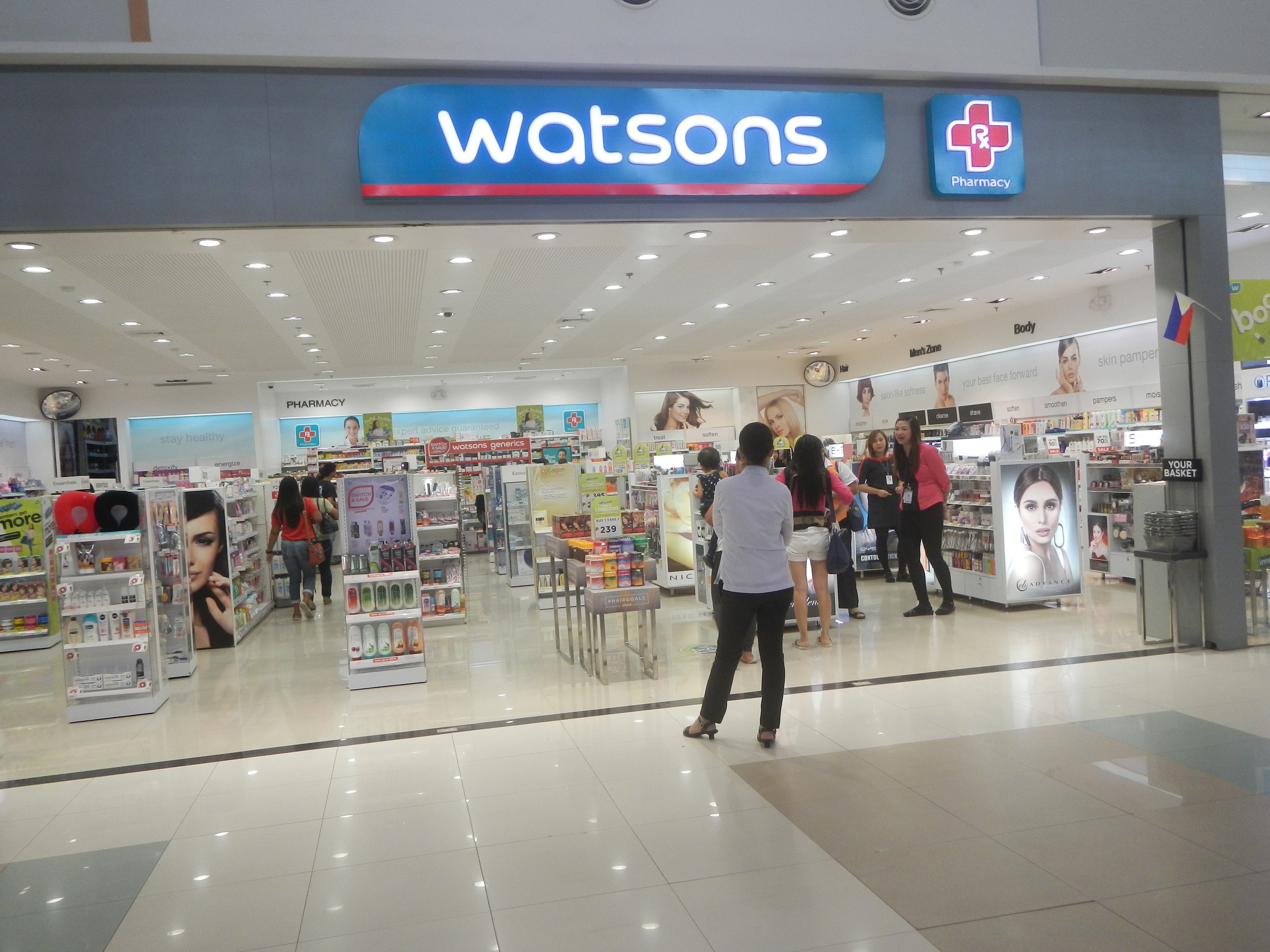 Pharmacy store interior in a shopping mall with customers, product displays, informational signs, a wall clock, and overhead lighting.
