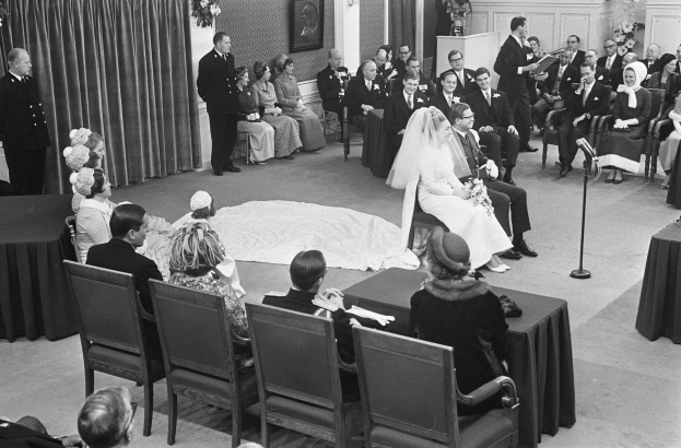 Schwarzes und weißes Foto einer Hochzeitszeremonie mit sitzenden Gästen, einem Mann mit Mikrofon, einer Frau mit Blumenstrauß, einem Podium mit Mikrofon und Blumenschmuck gegen eine Vorhangwand mit Rahmen und Tür.