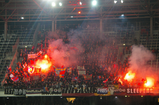 Eine große Menge Menschen in einem Stadion hält Fahnen und Banner, mit Leuchtspuren und Rauch, der aus metallenen Rahmen und Stangen aufsteigt und eine Decke mit Leuchten trägt.