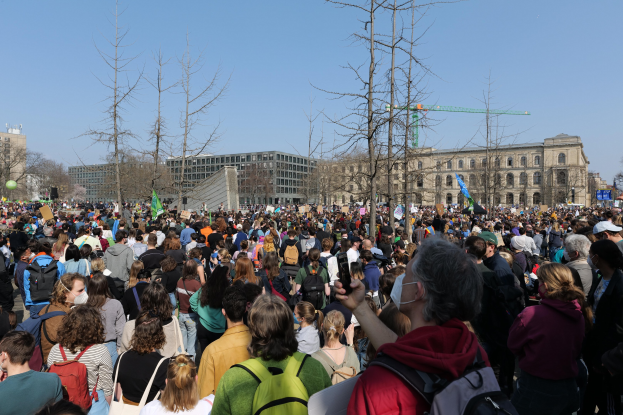 Eine große Menge Menschen nimmt an einer Klimawandel-Demonstration teil, hält Schilder und trägt Taschen, während sie vor einem Gebäude mit Fenstern und Bäumen unter einem klaren Himmel steht.