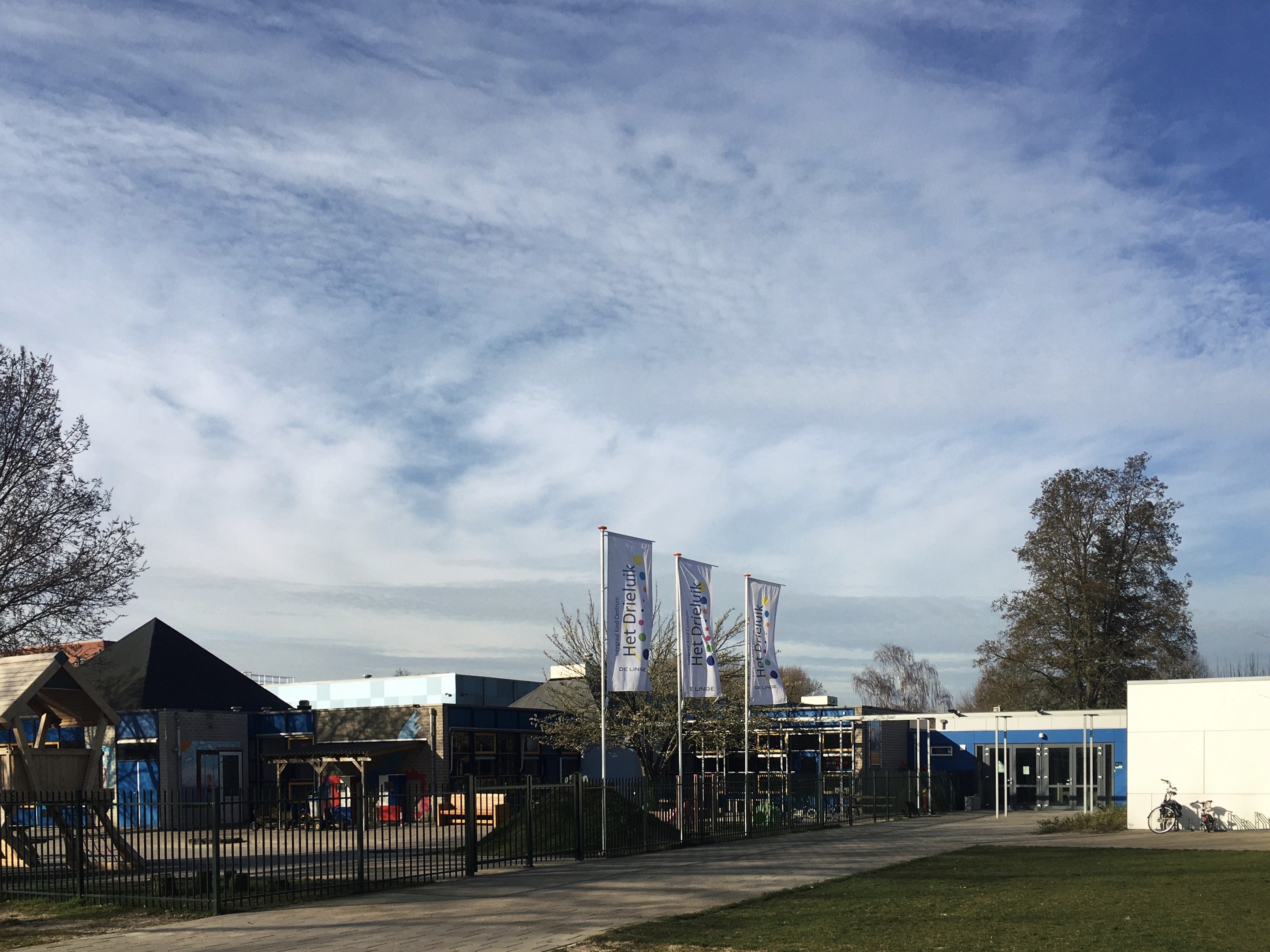 Großes Gebäude mit Metallzaun, Rasenfläche, Weg, Bäumen, Bannern, einem abgestellten Fahrrad, näheren Häusern und einem bewölktem Himmel, identifiziert als der neue Spielbereich für Kinder an der Schule.