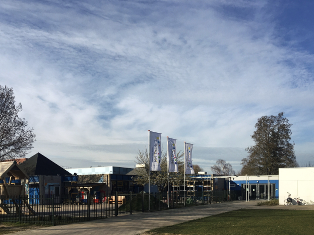 Großes Gebäude mit Metallzaun, Rasenfläche, Weg, Bäumen, Bannern, einem abgestellten Fahrrad, näheren Häusern und einem bewölktem Himmel, identifiziert als der neue Spielbereich für Kinder an der Schule.