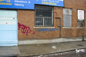 Eine Straßenszene mit einem Gehweg, einer verschmutzten Straße, einer Backsteinwand, Fenstern, Rohren, einem Tor und einer Werbetafel oben.