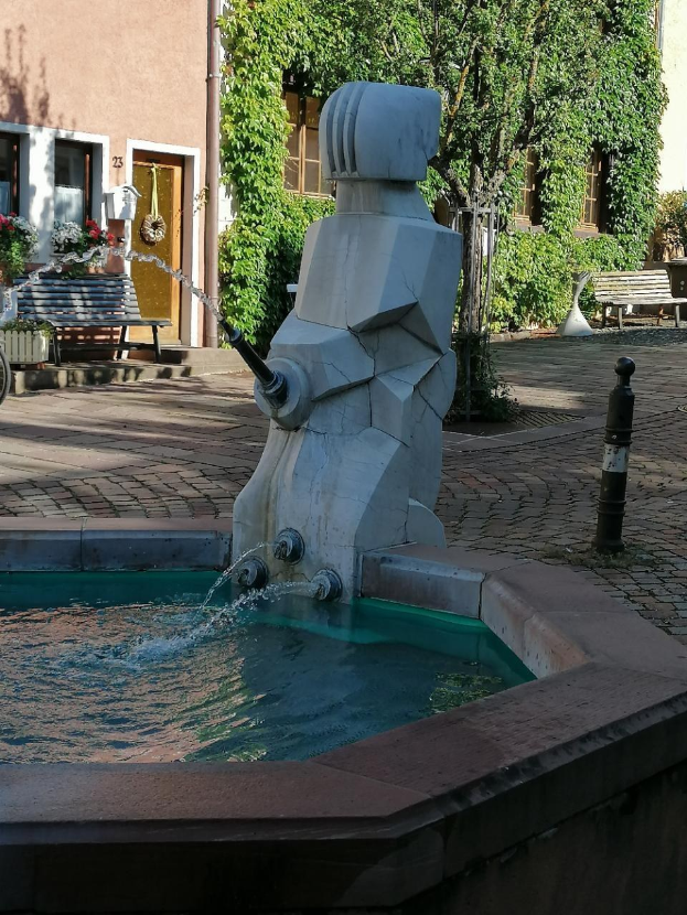 Ein steinerner Brunnen mit Wasserstrahlen in der Mitte eines Stadtplatzes, umgeben von Gebäuden, Bäumen, Bänken und Blumenkübeln.
