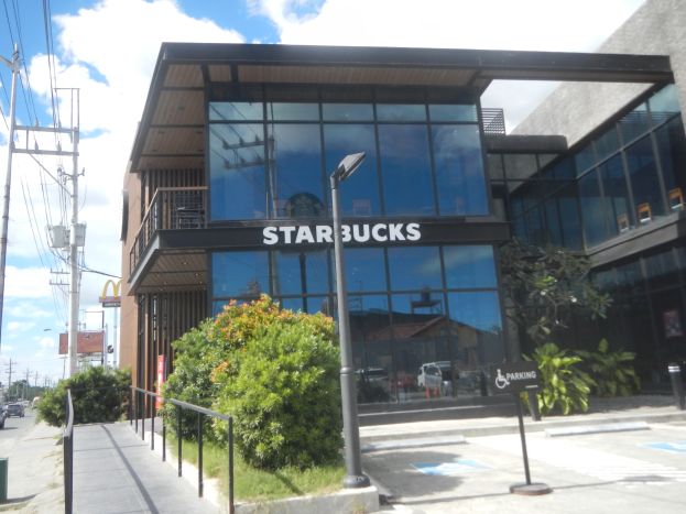 Außenansicht eines Starbucks-Cafés an einer Straßenkreuzung mit umgebender städtischer Infrastruktur und Fahrzeugen.
