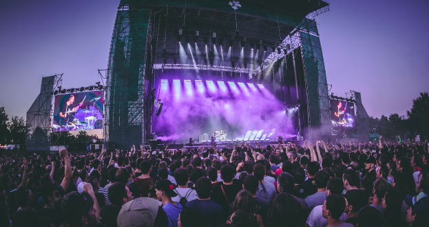 Eine große Menschenmenge auf einem Musikfestival, mit einer Bühne in der Mitte, auf der Musiker Instrumente spielen, beleuchtet von Scheinwerfern und umgeben von Bildschirmen, unter einem klaren blauen Himmel mit einem Gebäude und Bäumen im Hintergrund.