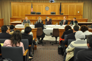 Ein Versammlungsraum mit einer Gruppe von Menschen, die auf St├╝hlen sitzen, zwei ausl├Ąndische Flaggen, ein orangefarbener Tisch mit Wasserflaschen und einem Mikrofon.