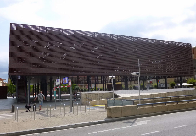 Großes Gebäude mit zahlreichen Fenstern, das als neuer Eingang zur Innenstadt dient, umgeben von Straßeninfrastruktur, Fahrzeugen, Fußgängern und Bäumen unter einem bewölkten Himmel.
