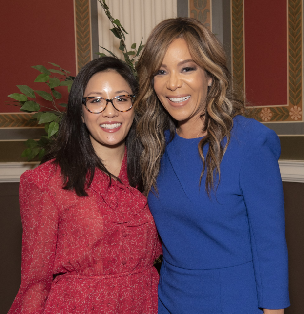 Zwei Frauen in Kleidern stehen strahlend nebeneinander auf der 2019 Women in Film Pre-Oscar-Luncheon, mit einer Pflanze und einem Vorhang im Hintergrund.