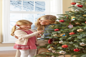 Ein geschmückter Weihnachtsbaum steht in einem Haus mit einer Frau und einem Kind im Vordergrund, einem Fenster und einer Wand im Hintergrund und Bäumen, die durch das Fenster sichtbar sind.