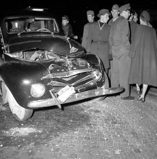 Schwarzes und wei?es Bild eines schwer beschdigten Autos am Stra?enrand mit einer eingedellten Haube und zersplitterten Fenstern, umgeben von einer Gruppe von Menschen, die die Szenerie begutachten.