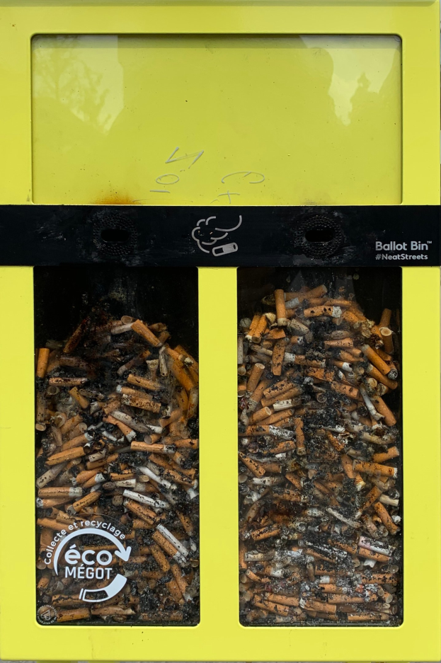 Gelber Mülltonne voller Zigarettenstummeln auf einem Gehweg, beschriftet für Zigarettenentfernung.