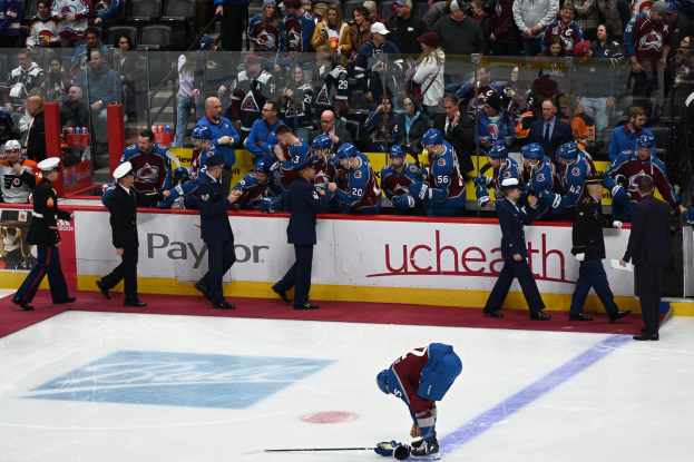 Ein Hockey-Spieler in dunkler Uniform steht auf dem Eis umgeben von Fans und Team-Mitarbeitern, mit Zuschauern hinter den Boards mit einem Spiel-Titel.