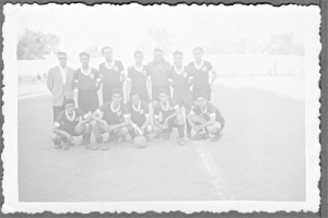 Ein Schwarz-Weiß-Foto in einem Rahmen, das fünf Männer in einer Mannschaftsposition mit einem Ball davor zeigt, sieben Männer dahinter, Sand unten, Bäume und eine Wand im Hintergrund und den Himmel oben.