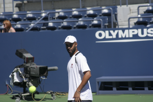 Ein Tennis-Spieler in Aktion mit einem Ball in der Luft, einer Kamera und einem Zaun im Hintergrund und einer Frau auf den Tribünen im Hintergrund.