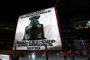 Eine nächtliche Stadionszene mit einem großen Banner in der Mitte, Zuschauern im Hintergrund, einem Glas auf dem Boden und einigen Spielern auf dem Feld, beleuchtet von Stadionlampen.