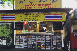 Ein Schaufenster mit verschiedenen Artikeln, darunter Getränkedosen, Speisekarten, eine Uhr, Gewürze in Plastikbehältern, Gewürze, Werbetafeln und Namensschilder, unter einem Himmel.