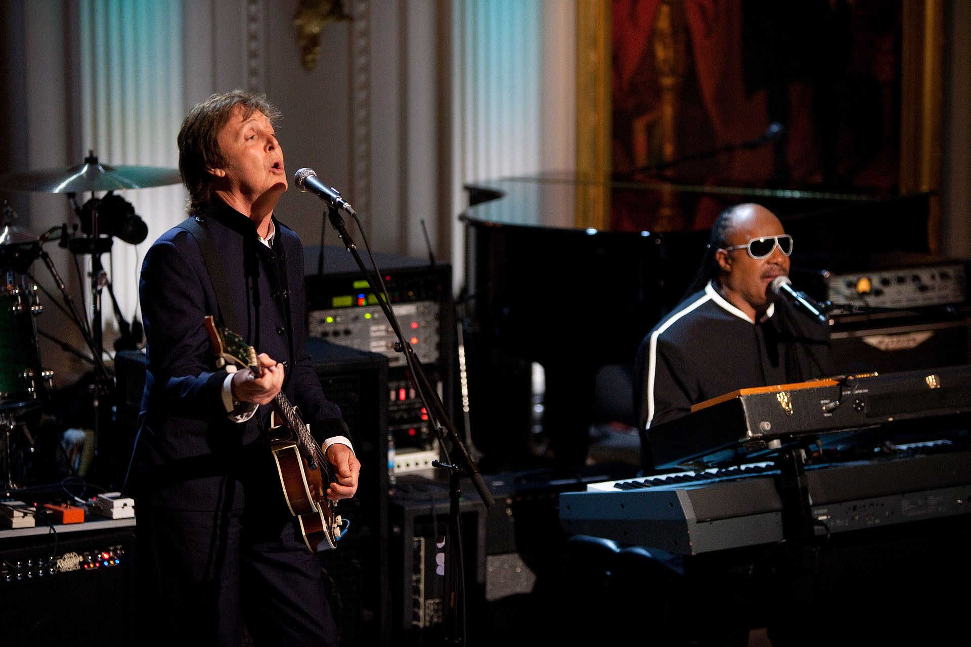 Paul McCartney spielt Gitarre und singt in ein Mikrofon, während Stevie Wonder, der eine Schutzbrille trägt, am Piano auf der Bühne steht, mit Musikinstrumenten und Dekoration im Hintergrund.