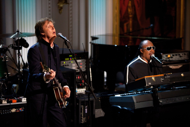 Paul McCartney spielt Gitarre und singt in ein Mikrofon, während Stevie Wonder, der eine Schutzbrille trägt, am Piano auf der Bühne steht, mit Musikinstrumenten und Dekoration im Hintergrund.