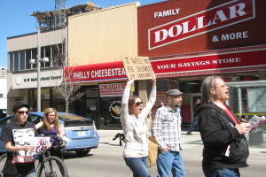 Eine Gruppe von Menschen demonstriert auf einer Straße mit Gebäuden, einem Pfahl, einem Baum und dem Himmel im Hintergrund, wobei ein Auto und ein Fahrrad auf der linken Seite zu sehen sind.