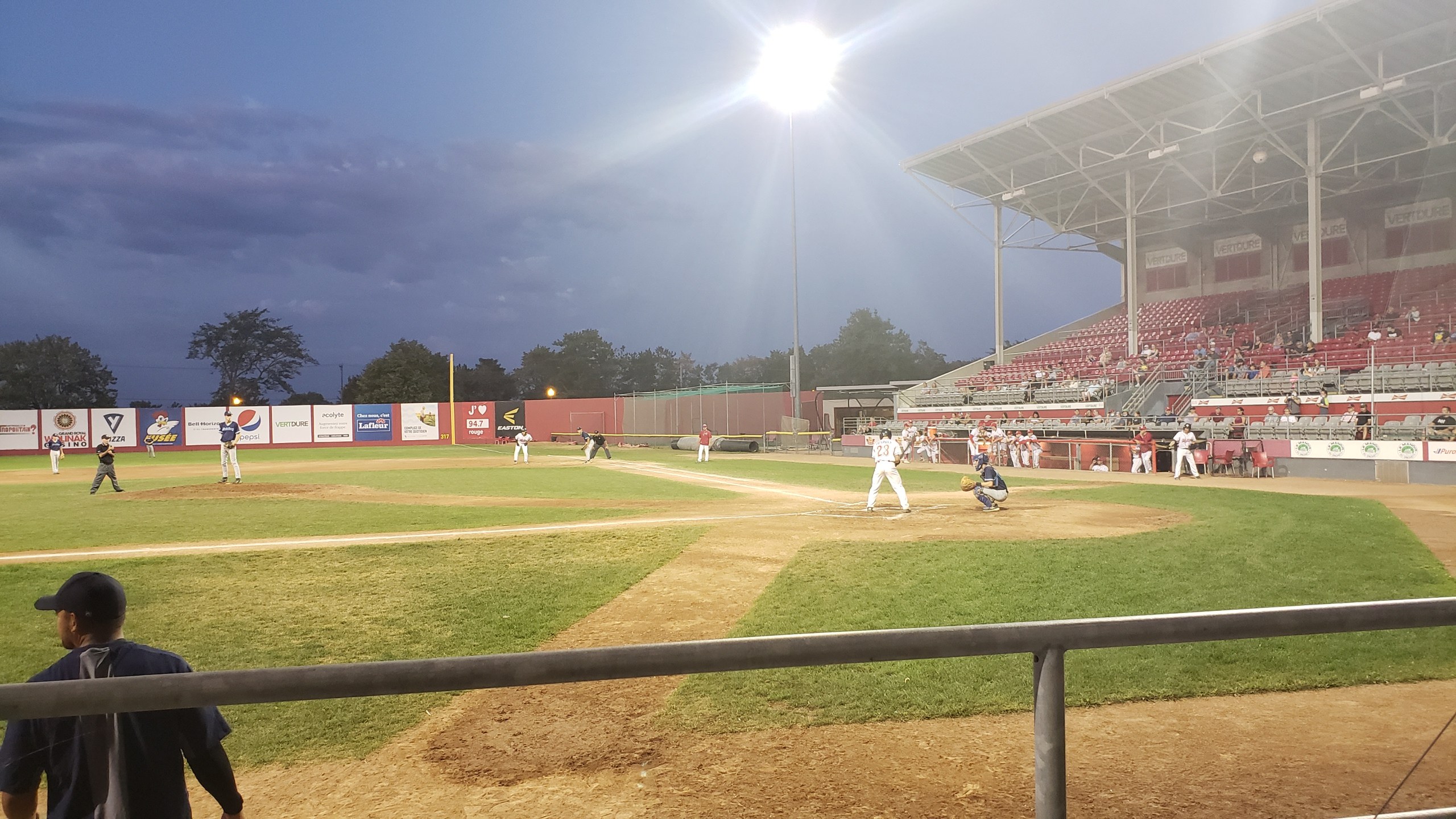 Baseballspiel in einem Stadion mit Zuschauern auf den RÀngen, BÀumen, MÀsten, Lichtern, Werbetafeln und einem klaren blauen Himmel im Hintergrund.
