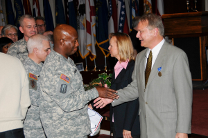 Eine Gruppe von Menschen, die mit zwei Personen Handschlag machen, Flaggen im Hintergrund an Stangen, ein Podium mit Mikrofon und eine Topfpflanze in der Nähe.