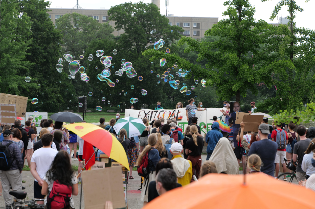 Eine Gruppe von Menschen vor einer Protestmenge mit Schirmen, Taschen und Schildern, mit Seifenblasen in der Luft, Bäumen, Gebäuden, einem Turm und einem klaren blauen Himmel im Hintergrund.