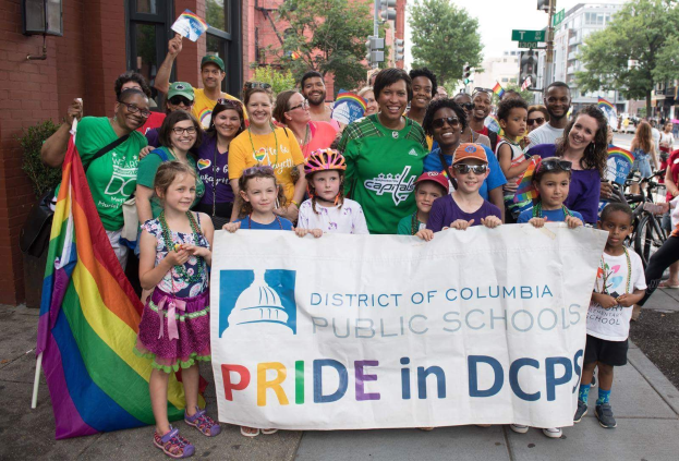 Gruppe von Menschen auf dem Boden, einige halten ein Banner mit der Aufschrift "Bezirksschulen Washington DC Pride in DCPS", mit Kindern, die eine Fahne halten, einem Radfahrer, Bäumen, Gebäuden mit Fenstern, Schildern, Straßenlaternen, Ampeln und einem bewölkten Himmel.