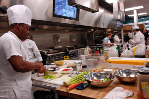 Eine Gruppe von Köchen in einer Küche beim Zubereiten von Speisen, mit einem Koch im Vordergrund in einer weißen Schürze und Mütze, der ein Messer hält und von verschiedenen Utensilien, Schüsseln, Gemüse, einem Handy, Flaschen und anderen Gegenständen umgeben ist, mit einem Fernseher und Deckenleuchten im Hintergrund.