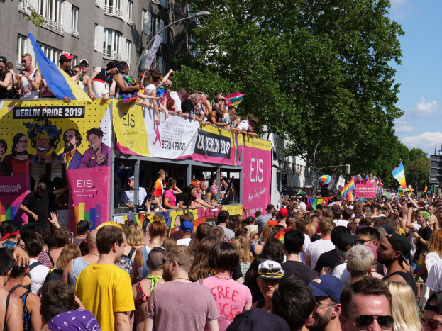 Eine große Menge marschiert während des Berlin Pride 2019 die Straße entlang, viele tragen Mützen und Schutzbrillen, einige halten Fahnen, mit Bannern, Laternenmasten, Bäumen und Gebäuden im Hintergrund bei bewölktem Himmel.