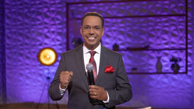 Stephen Colbert, Moderator von *The Daily Show with Trevor Noah*, steht auf einer Bühne und hält ein Mikrofon in der Hand, trägt einen Anzug, Krawatte und Brille, mit sichtbarer Beleuchtungsausrüstung und Gegenständen hinter ihm.