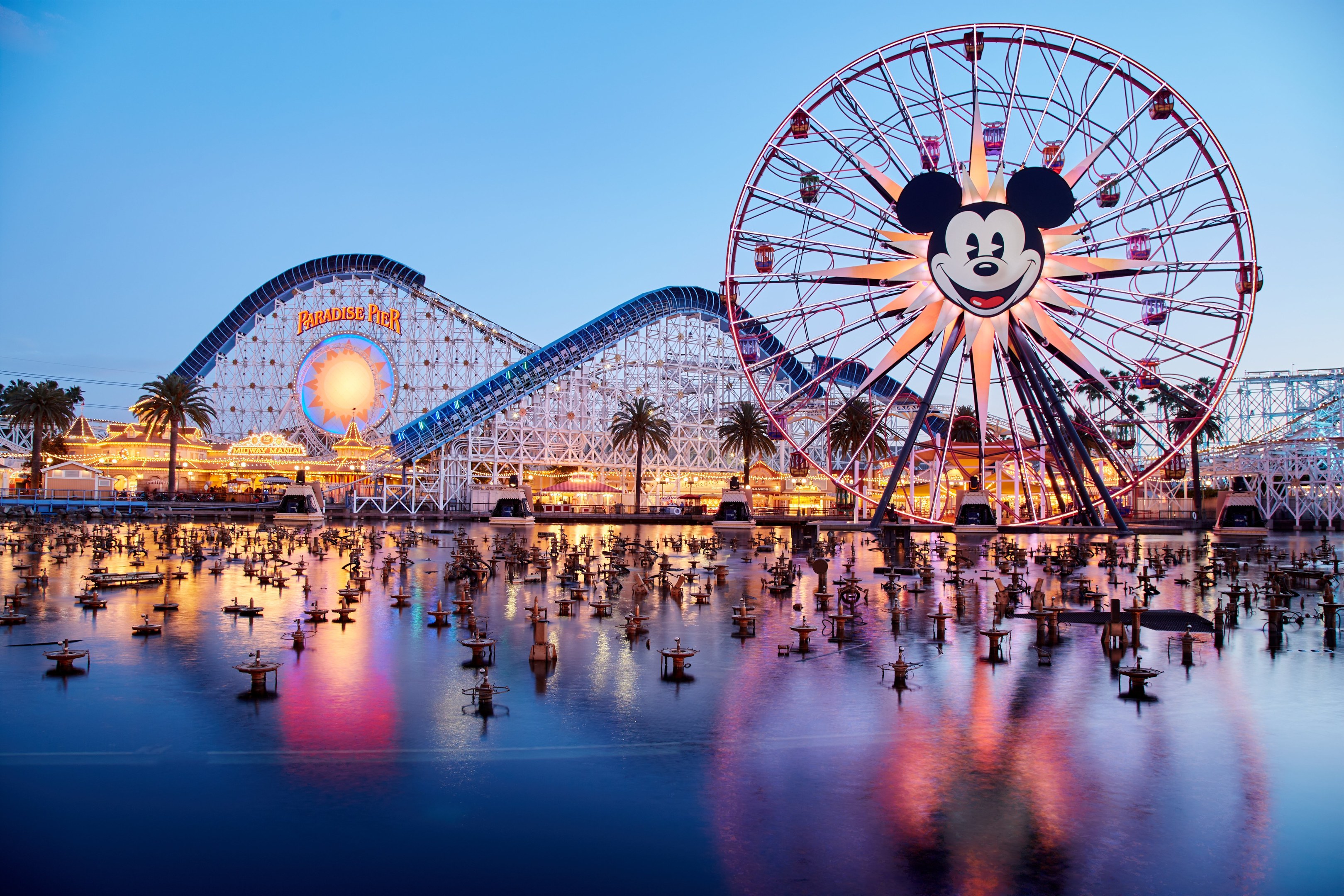 Eine lebendige Dämmerungsszene im Disneyland California Adventure mit einem riesigen Mickey-Maus-Rad im Vordergrund, einer Achterbahn im Hintergrund und Booten auf dem Wasser, mit einem Himmel aus Orangen, Pink- und Purpurfarben und silhouettenartigen Bäumen.